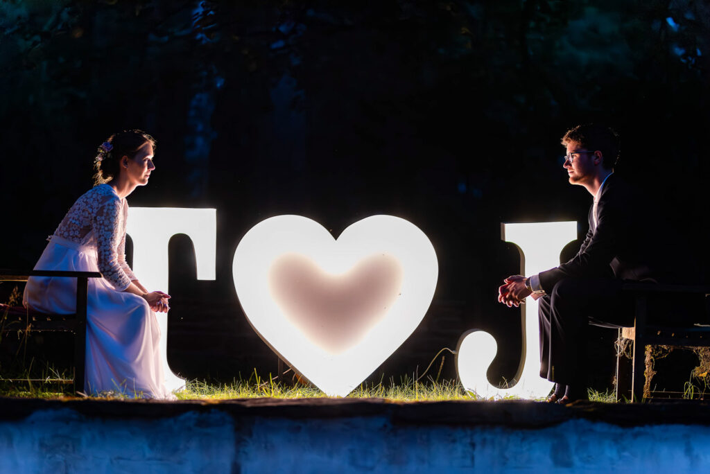 Nevěsta a ženich sedící naproti sobě u svítící dekorace s nápisem „T ❤️ J“ ve večerním světle