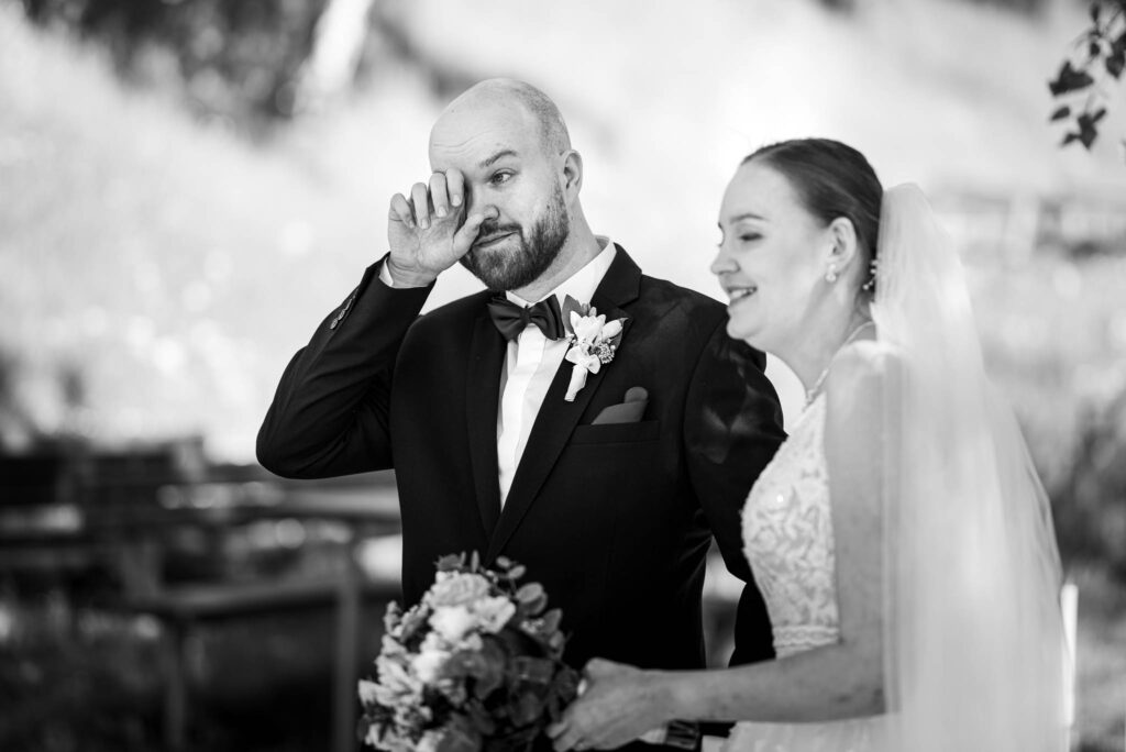 Černobílá fotografie ženicha, který si utírá slzu, stojící vedle usmívající se nevěsty.