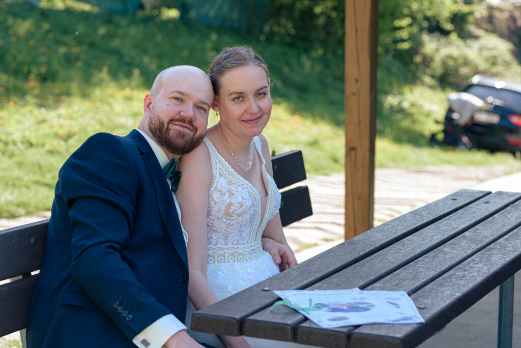 Nevěsta a ženich sedí na lavičce, opřeni o sebe, a usmívají se do objektivu.